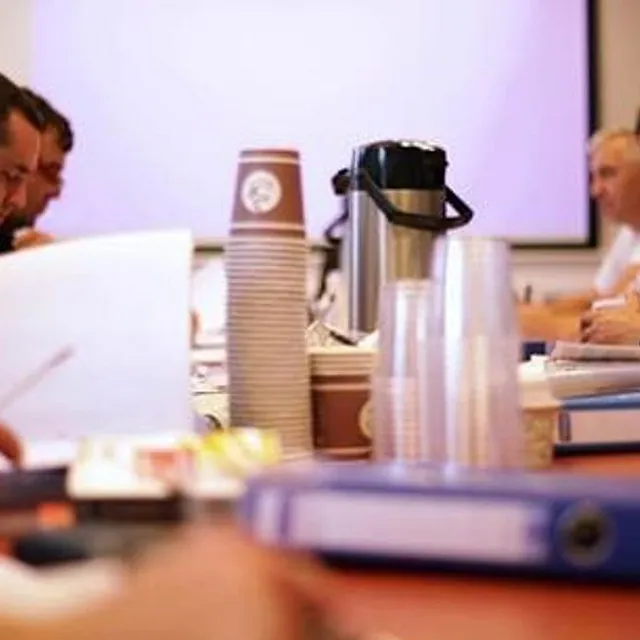 Gruppe voksne kursdeltakere sitter rundt bord med permer og kaffekopper.