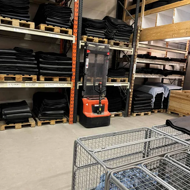 Lager med paller av matter, traller og en palletruck.