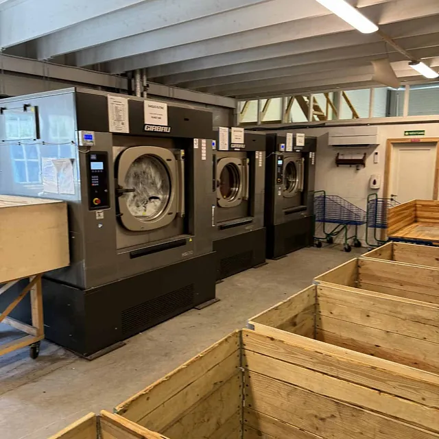 Tre store industrivaskemaskiner står på rekke i et vaskeri, foran flere tomme trekasser.